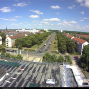 Das Foto zeigt eine weite Stadtansicht von einem Dach aus, mit einer breiten, von Bäumen gesäumten Straße und mehreren Wohn- und Geschäftsgebäuden. Am Himmel sind viele kleine Wolken zu sehen, während unten Autos auf den Straßen fahren.