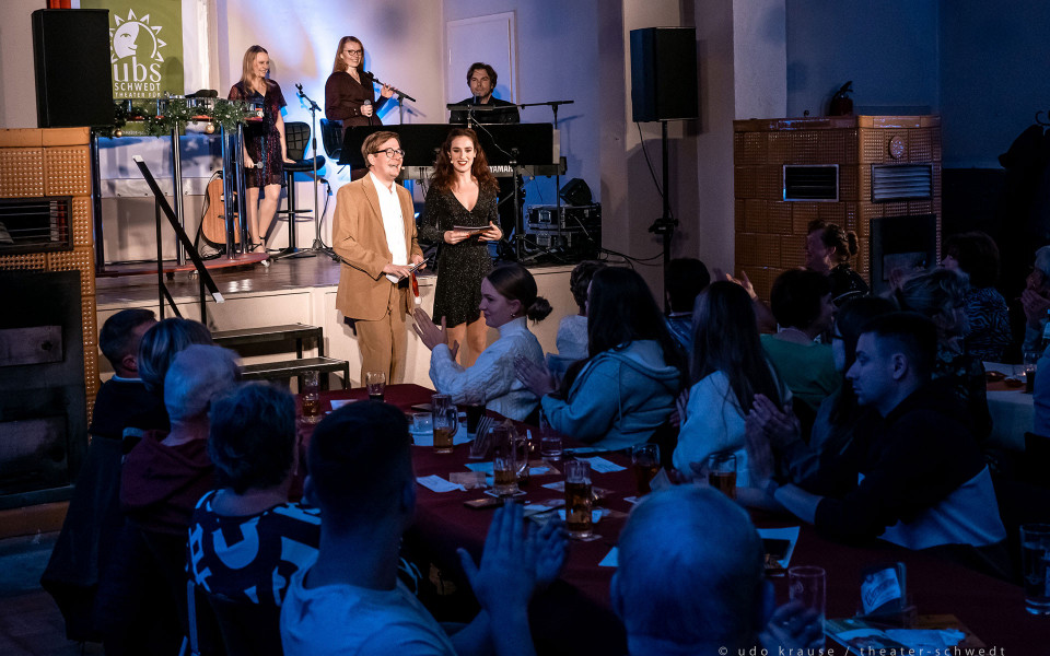 Auf einer kleinen Bühne stehen mehrere Künstlerinnen und ein Pianist, während zwei Moderatoren vor dem Publikum lächeln. Die Gäste sitzen an Tischen mit Getränken und applaudieren in einer stimmungsvoll beleuchteten Theater- oder Veranstaltungsatmosphäre.