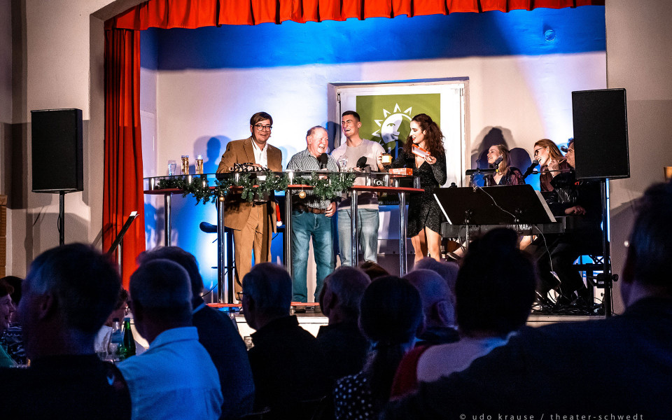 Auf einer kleinen Bühne mit rotem Vorhang stehen mehrere Personen vor Publikum und lachen, während Mikrofone, Gläser und festliche Dekoration auf den Stehtischen zu sehen sind. Rechts begleitet eine Musikgruppe mit Keyboard und Mikrofonen die Veranstaltung, die von blauem Bühnenlicht beleuchtet wird.