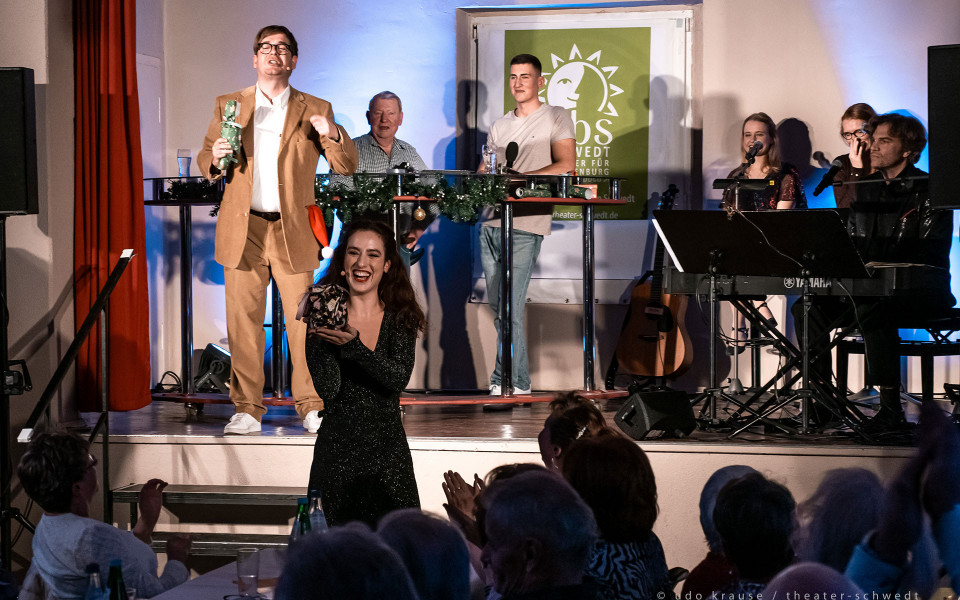 Auf einer kleinen Bühne treten mehrere Personen bei einer festlichen Veranstaltung auf, während vorne eine lächelnde Frau in einem glitzernden Kleid ein Geschenk präsentiert. Rechts begleiten Musiker am Keyboard und mit Mikrofonen die Szene, und im Publikum wird applaudiert.
