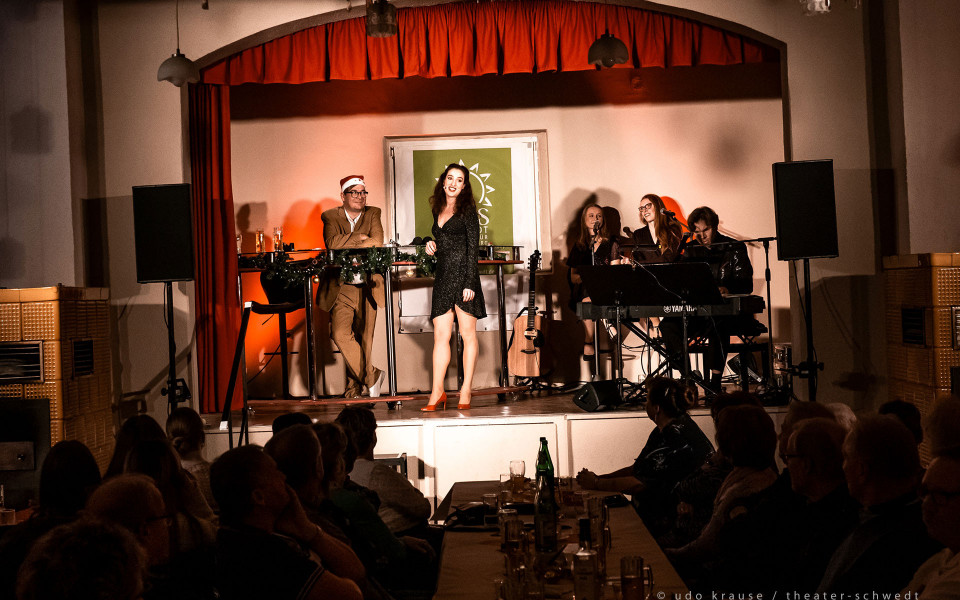 Auf einer kleinen Bühne mit rotem Vorhang steht eine Sängerin im schwarzen Kleid im Scheinwerferlicht, während neben ihr ein Mann mit Weihnachtsmütze an einem Stehtisch lehnt. Rechts begleiten mehrere Musikerinnen und ein Pianist die Aufführung, vor ihnen sitzt ein Publikum an Tischen im abgedunkelten Saal.