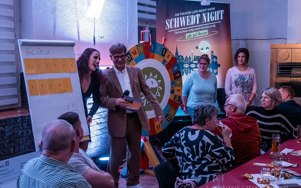 Bei einer Theater-Late-Night-Show stehen ein Moderator mit Mikrofon und mehrere Teilnehmer vor einem Glücksrad und einer Tafel mit nummerierten Karten. Das Publikum sitzt an Tischen mit Getränken und schaut der Szene in einem gemütlich beleuchteten Veranstaltungsraum zu.