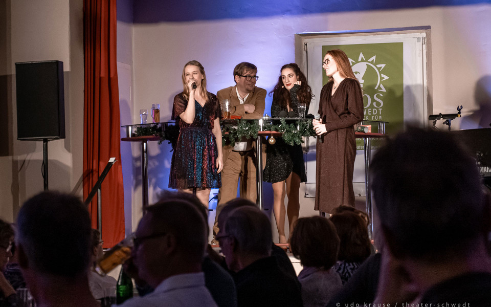 Auf einer Bühne stehen vier Personen vor einem Publikum, zwei Frauen halten Mikrofone und wirken, als würden sie sprechen oder singen. Die Szene ist festlich beleuchtet, mit Stehtischen, Getränken, Dekoration und einem grünen Plakat im Hintergrund.
