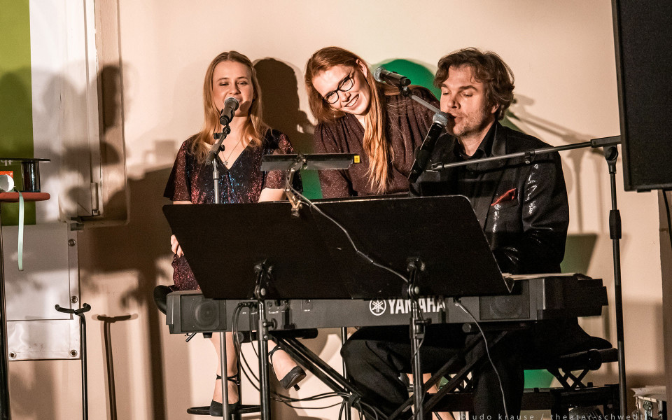 Drei Musiker sitzen auf einer kleinen Bühne vor Mikrofonen, während ein Mann am Yamaha-Keyboard spielt. Die beiden Frauen neben ihm lächeln und singen, beleuchtet von warmem Bühnenlicht.
