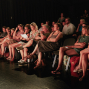 Ein Publikum sitzt in einem dunklen Theaterraum auf Stühlen und schaut aufmerksam nach vorne zur Bühne. Warmes Licht beleuchtet die Gesichter der Erwachsenen und Kinder, während der Hintergrund fast vollständig schwarz bleibt.