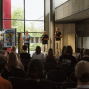 Drei Männer stehen mit Mikrofonen vor einem sitzenden Publikum in einer modernen Halle und moderieren offenbar eine Veranstaltung. Neben ihnen sind Plakate und Dekorationen zum Schul-Theater-Fest 2026 zu sehen.