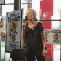 Eine Frau steht bei einer Veranstaltung in einem hellen Foyer und spricht mit einem Mikrofon vor Plakaten eines Schultheater-Festivals. Neben ihr sind Luftballons, ein Mikrofonständer und große Fenster mit Blick ins Grüne zu sehen.