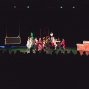 Auf einer dunklen Theaterbühne führen mehrere Kinder in bunten Kostümen eine Szene auf, während links eine hängende Requisite und rechts ein bemaltes Bühnenelement zu sehen sind. Im Vordergrund sitzen die Zuschauer als dunkle Silhouetten, während die Bühne von wenigen grünen Scheinwerfern beleuchtet wird.