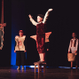 Auf einer dunklen Theaterbühne steht eine junge Darstellerin in einem roten Kostüm auf einem Hocker und hebt die Arme. Um sie herum stehen weitere kostümierte Personen, während links eine hängende Requisite aus Seilen zu sehen ist.