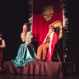 Kinder führen auf einer Bühne ein Theaterstück in historischen Kostümen auf, mit einer Prinzessin im türkisfarbenen Kleid und einem Jungen im roten Umhang auf einem erhöhten Podest. Im Hintergrund hängt ein roter Vorhang mit Wappen, während weitere Kinder mit roten Wangen geschminkt am Rand der Szene stehen.