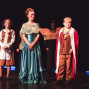 Drei Kinder stehen in fantasievollen Kostümen auf einer dunkel beleuchteten Theaterbühne. Links sind Seile und Bühnenelemente zu sehen, während im Hintergrund weitere verkleidete Personen im Schatten stehen.