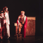 Auf einer dunklen Theaterbühne stehen mehrere Kinder in fantasievollen Kostümen, darunter eine Hexe, ein Junge in rotem Samt und eine Figur in einem weißen Kleid. Rechts sitzt ein blondes Kind mit rotem Umhang auf einem Stuhl vor einer bemalten Kulisse und blickt zu der Gruppe hinüber.