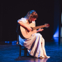 Eine junge Frau sitzt auf einem Hocker auf einer dunklen Bühne und spielt konzentriert eine klassische Gitarre. Blaues und warmes Bühnenlicht beleuchten sie und ihr langes helles Kleid vor einem fast schwarzen Hintergrund.