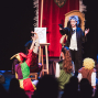 Kinder führen auf einer Bühne ein Theaterstück auf, verkleidet als Zauberer, Narr und andere Figuren. Ein Kind mit blauer Perücke steht vor einem großen roten Vorhang neben einer Staffelei, während die anderen Kinder zu ihm hinaufschauen und die Hände heben.