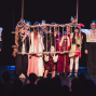 Auf einer Theaterbühne stehen mehrere Kinder und Jugendliche in fantasievollen Kostümen um einen hängenden Käfig mit Seilen. Das dunkle Bühnenlicht und die sichtbaren Zuschauer im Vordergrund lassen die Szene wie eine dramatische Aufführung wirken.
