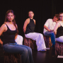 Mehrere Jugendliche sitzen auf einer dunklen Bühne auf hölzernen Hockern und blicken ernst in verschiedene Richtungen. Die Szene ist warm und farbig beleuchtet, während der schwarze Hintergrund den Eindruck einer Theaterprobe oder Aufführung verstärkt.
