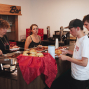 An einer Bar werden mehrere Personen mit warmem Essen wie Würstchen und Brötchen bedient. Hinter der Theke stehen Mitarbeiterinnen, während Gäste mit Tellern in einer Schlange warten.