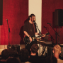 Ein Musiker steht auf einer kleinen Bühne, singt in ein Mikrofon und spielt eine schwarze E-Gitarre. Vor ihm schaut ein Publikum zu, während rote Wandpaneele, Lautsprecher und weiteres Bühnenequipment den Raum prägen.