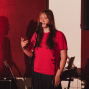 Eine junge Frau in einem roten T-Shirt steht auf einer Bühne vor einem Mikrofon und spricht oder singt. Im Hintergrund sind rote Vorhänge, Musikinstrumente und ein großer Lautsprecher zu sehen.