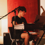 Ein junger Musiker sitzt an einem elektronischen Keyboard und schaut konzentriert auf die Noten oder ein Display. Um ihn herum stehen Mikrofone und Kabel, während warmes Licht vor einem roten Vorhang eine intime Studioatmosphäre schafft.