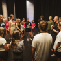 Eine Gruppe von Jugendlichen und Erwachsenen steht im Kreis auf einer Bühne vor schwarzen Vorhängen. Ein Mann spricht zur Gruppe, die aufmerksam zuhört, als ob sie an einer Theaterprobe oder einem Workshop teilnimmt.