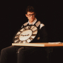 Ein junger Mann mit Brille sitzt auf einer dunklen Bühne und hält eine große runde Uhr vor sich. Vor ihm steht ein Tisch mit einem Glas und weiteren Gegenständen, während warmes Licht die Szene beleuchtet.