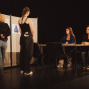 Auf einer dunklen Bühne stehen zwei Personen vor einer Stellwand mit Plakaten, während zwei junge Frauen an einem Tisch sitzen und ihnen gegenüberblicken. Die Szene wirkt wie eine Beratung, Prüfung oder Theaterprobe mit schlichtem Bühnenaufbau und gedämpftem Licht.