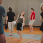 Mehrere junge Menschen bewegen sich barfuß in einer Turnhalle, als würden sie an einer Tanz- oder Theaterübung teilnehmen. In der Mitte steht eine Person mit einem Blatt Papier in der Hand und blickt zu den anderen Teilnehmenden.