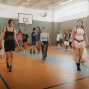 In einer hellen Sporthalle mit Basketballkorb laufen mehrere junge Menschen locker umher, teils barfuß oder in Socken. Im Vordergrund gehen drei Personen in auffälliger Freizeitkleidung auf die Kamera zu, während im Hintergrund weitere Gruppen miteinander interagieren.