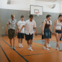 Eine Gruppe junger Menschen bewegt sich in einer Turnhalle über den orangefarbenen Sportboden, einige barfuß oder in Socken. Im Hintergrund sind ein Basketballkorb, graue Wände und weitere Personen zu sehen.