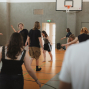 Mehrere Personen bewegen sich in einer Sporthalle, vermutlich bei einer Tanz- oder Bewegungsübung. Im Hintergrund sind ein Basketballkorb, Kletterstangen und eine geöffnete Tür zu sehen.