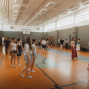 In einer hellen Sporthalle stehen und gehen viele junge Menschen in lockerer Kleidung und Socken auf dem orangefarbenen Hallenboden. An den Wänden sind Bänke, Sportgeräte und ein Basketballkorb zu sehen, während die Gruppe offenbar an einer gemeinsamen Übung oder Probe teilnimmt.