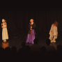 Auf einer dunklen Theaterbühne stehen drei kostümierte Kinder vor einem schwarzen Vorhang. Im Vordergrund sind die Silhouetten des Publikums zu sehen, während die Darsteller von warmem Bühnenlicht beleuchtet werden.