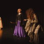 Auf einer dunklen Theaterbühne stehen drei kostümierte Personen im Scheinwerferlicht vor einem schwarzen Vorhang. In der Mitte trägt eine Figur einen schwarzen Mantel und einen violetten Rock, während rechts eine zerlumpte Gestalt mit langen Haaren und links eine Person in einem hellen Kleid zu sehen ist.