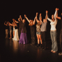 Eine Gruppe von Darstellerinnen und Darstellern steht auf einer dunklen Theaterbühne in einer Reihe und hält sich mit erhobenen Armen an den Händen. Warmes Bühnenlicht beleuchtet die unterschiedlich gekleideten Personen, als würden sie sich nach einer Aufführung verbeugen oder vom Publikum feiern lassen.