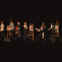 Auf einer dunklen Bühne stehen mehrere Menschen zwischen Staffeleien mit bunten Gemälden und wirken wie bei einer Kunst- oder Theateraufführung. Im Vordergrund sind die Silhouetten des Publikums zu sehen, während die Darstellerinnen und Darsteller miteinander interagieren.