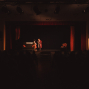 Auf einer dunkel beleuchteten Theaterbühne stehen zwei Schauspieler im Scheinwerferlicht vor einem roten Vorhang. Im Vordergrund sind die Zuschauer nur als dunkle Silhouetten zu erkennen, während auf der Bühne ein Sofa und ein Sessel als Kulisse stehen.