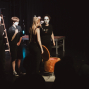 Auf einer dunklen Theaterbühne stehen mehrere schwarz gekleidete Personen mit weiß geschminkten Gesichtern im Scheinwerferlicht. Im Vordergrund sind Publikumssilhouetten zu sehen, während auf der Bühne ein orangefarbener Sessel, eine Leiter und eine Gitarre im Hintergrund stehen.