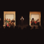 Auf einer dunklen Theaterbühne sind zwei beleuchtete, wie Zugabteile wirkende Sitzbereiche mit mehreren Darstellern zu sehen. Im Vordergrund erkennt man die Silhouetten des Publikums, während weitere Schauspieler in Uniformen seitlich und in der Mitte stehen.