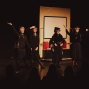 Auf einer Theaterbühne stehen vier schwarz gekleidete Darstellerinnen in eleganten Kostümen und posieren mit erhobenen Armen vor einem Bühnenbild, das wie ein Zugabteil wirkt. Im Vordergrund sind die dunklen Silhouetten des Publikums zu sehen, während warmes Licht die Szene beleuchtet.