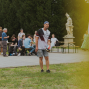 Ein Mann steht auf einer Wiese und hält einen Volleyball in der Hand, während im Hintergrund mehrere Personen an Biertischen sitzen oder stehen. Hinter ihnen sind ein Wald, eine Statue und ein gelblicher unscharfer Vordergrund zu sehen.