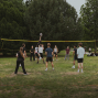 Eine Gruppe Jugendlicher spielt auf einer Wiese im Park Volleyball an einem gelben Netz. Im Hintergrund stehen Bäume und weitere Zuschauer sitzen oder stehen am Spielfeldrand.
