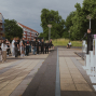 Eine Gruppe von Menschen steht auf einem gepflasterten Platz und hört einer Person zu, die auf einer kleinen Bühne an einem Mikrofon spricht. Im Hintergrund sind Wohnhäuser, Bäume und ein bewölkter Himmel zu sehen, während rechts weiße Ausstellungsobjekte und ein transparenter Pavillon stehen.