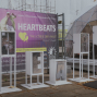 Vor einem großen violetten Werbebanner für „HEARTBEATS“ stehen weiße, beleuchtete Buchstabeninstallationen, die „HEART“ bilden. Rechts daneben befindet sich eine transparente Zelt- oder Bühnenkonstruktion auf einem gepflasterten Platz.