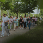 Eine größere Gruppe von Menschen geht an einem sonnigen Tag auf einem von Bäumen gesäumten Gehweg entlang. Viele tragen Namensschilder und wirken wie Teil einer gemeinsamen Führung oder Veranstaltung.