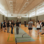 Eine Gruppe von Menschen steht in einer großen Sporthalle im Kreis und hört einer Person in der Mitte zu. Die Halle ist hell beleuchtet, hat hohe Fenster, einen Basketballkorb und markierte Spielfeldlinien auf dem Boden.