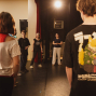 Eine Gruppe von Menschen steht auf einer Bühne oder in einem Proberaum und wirkt, als würde sie an einer Theater- oder Workshopübung teilnehmen. Im Vordergrund sind zwei Personen von hinten zu sehen, während in der Mitte mehrere Teilnehmende unter warmem Bühnenlicht stehen.