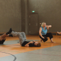 In einer Sporthalle liegen zwei Kinder spielerisch auf dem Boden und heben die Beine, während sie offenbar eine Übung machen. Daneben hocken zwei Frauen und unterhalten sich lachend, eine davon gestikuliert lebhaft.
