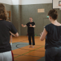 In einer Sporthalle leitet ein barfüßiger Mann in schwarzer Kleidung eine Gruppe bei ruhigen Bewegungsübungen an. Mehrere Teilnehmende stehen im Kreis und folgen seinen Handbewegungen, während im Hintergrund ein Basketballkorb zu sehen ist.