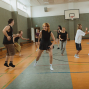 Mehrere junge Erwachsene bewegen sich lachend und spielerisch in einer Turnhalle mit Basketballkorb. Sie tragen Freizeit- oder Sportkleidung und scheinen gemeinsam zu tanzen oder eine Übung zu machen.